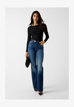 Haut noir à manches longues avec motif subtil, jean bleu taille haute, et pochette noire ; le modèle porte des talons noirs pointus.