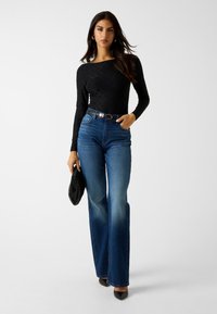 Top nero a maniche lunghe con motivo sottile, jeans blu a vita alta e pochette nera; il modello indossa tacchi a punta neri.