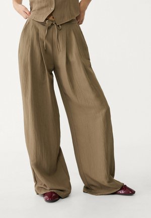 Personne portant un pantalon ample à taille haute de couleur marron avec une taille à cordon, associé à des chaussures pointues brillantes rouge foncé.