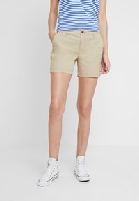Beige bomuldsshorts med opkrukkede kanter, forlommer og knaplukning, kombineret med en blå og hvid stribet tanktop og hvide sneakers.