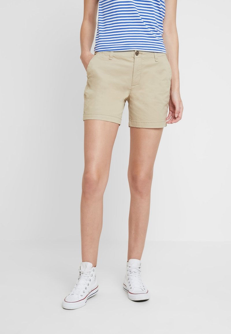 Beige bomuldsshorts med opkrukkede kanter, forlommer og knaplukning, kombineret med en blå og hvid stribet tanktop og hvide sneakers.