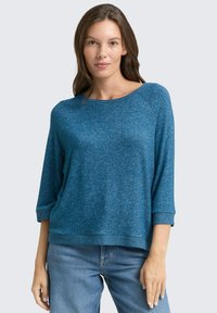 Blauer melierter Strickpullover mit weitem Ausschnitt, Dreiviertelärmeln und gerippten Bündchen. Kombiniert mit hellen Jeans.