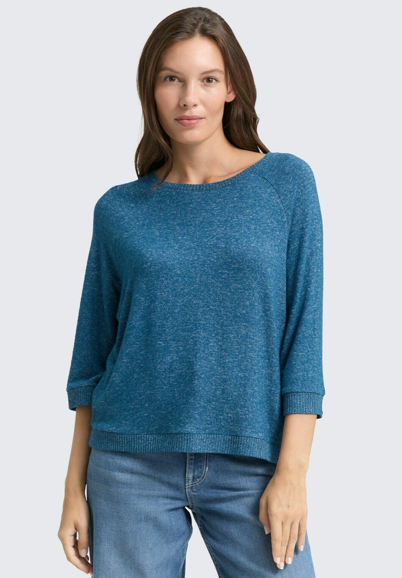Blauer melierter Strickpullover mit weitem Ausschnitt, Dreiviertelärmeln und gerippten Bündchen. Kombiniert mit hellen Jeans.