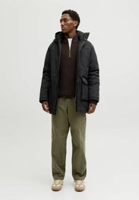 Giacca nera isolata con cappuccio, chiusure con zip e bottoni, indossata sopra un maglione a maglia marrone scuro, abbinata a pantaloni verde chiaro e sneakers beige.