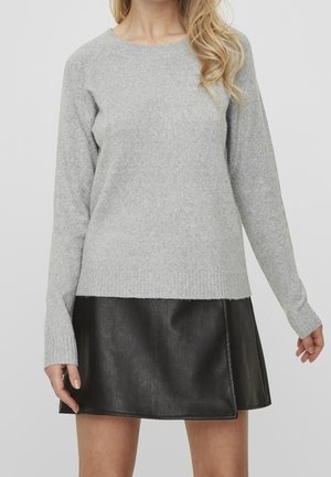 Frau trägt einen hellgrauen, langärmeligen Strickpullover und einen kurzen schwarzen Lederrock vor einem schlichten hellen Hintergrund.