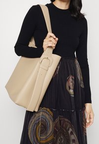Borsa tote in pelle beige con un design strutturato, dotata di una larga tracolla e un evidente dettaglio con fibbia. Gonna scura a fantasia visibile sotto.