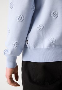 Pull en maille bleu clair avec motif en relief du logo, poignets et ourlet côtelés, avec une étiquette noire affichant le nom de la marque.