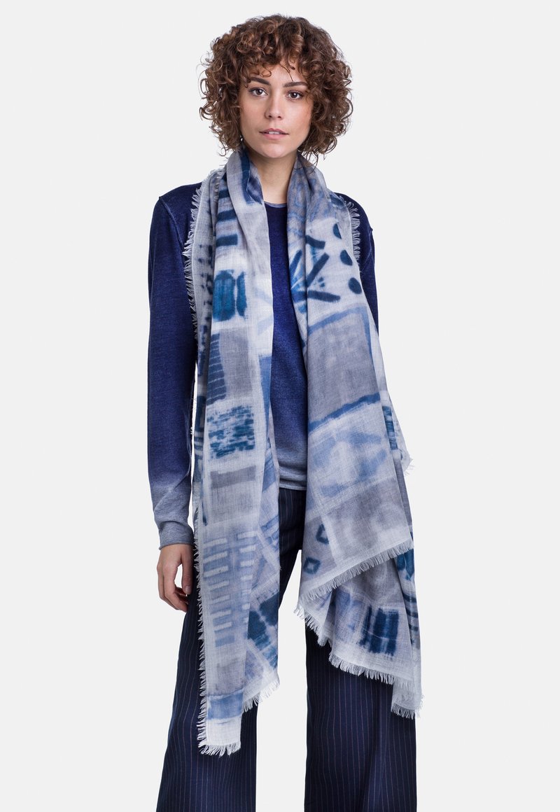 Sciarpa leggera grigia con motivi astratti blu; bordi frangiati; indossata sopra una maglia blu e abbinata a pantaloni a vita alta a righe.