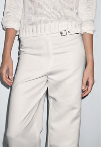 Witte gebreide trui met opgerolde mouwen, gecombineerd met hoge, crèmekleurige wijde broekspijpen, met een zichtbare riem met een zilveren gesp.