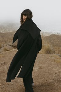 Woman with wavy hair in a long black coat walking on rocky terrain with dry grass, looking back over her shoulder toward the camera.