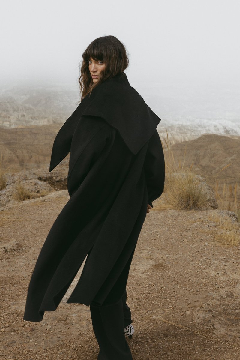 Woman with wavy hair in a long black coat walking on rocky terrain with dry grass, looking back over her shoulder toward the camera.