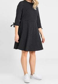 Černé šaty s puntíky, tříčtvrteční rukávy a mašle na rukávech, vyrobené z lehké látky, nošené s bílými teniskami.