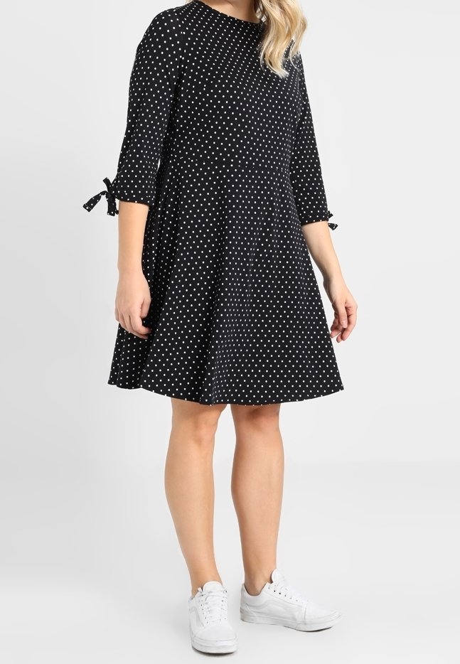 Černé šaty s puntíky, tříčtvrteční rukávy a mašle na rukávech, vyrobené z lehké látky, nošené s bílými teniskami.