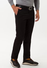 Pantaloni neri con vestibilità slim, realizzati in un tessuto liscio. Presentano tasche laterali, una cintura nera e sono indossati con sneaker nere e arancioni.