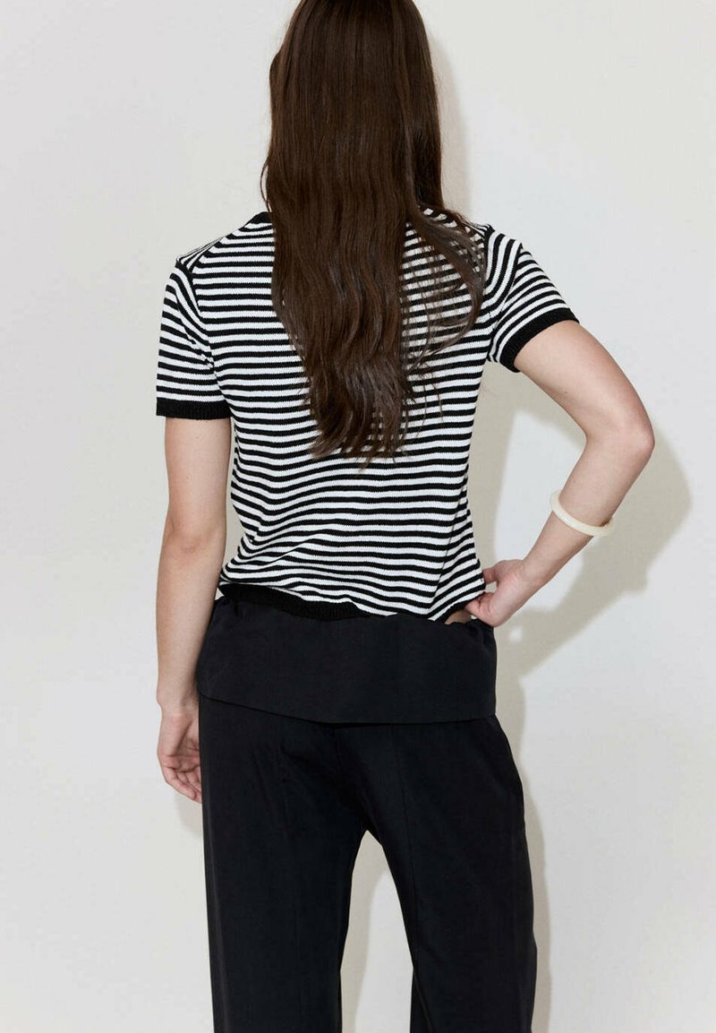 Femme aux longs cheveux bruns portant un haut à manches courtes rayé noir et blanc et un pantalon noir, debout avec une main sur la hanche.
