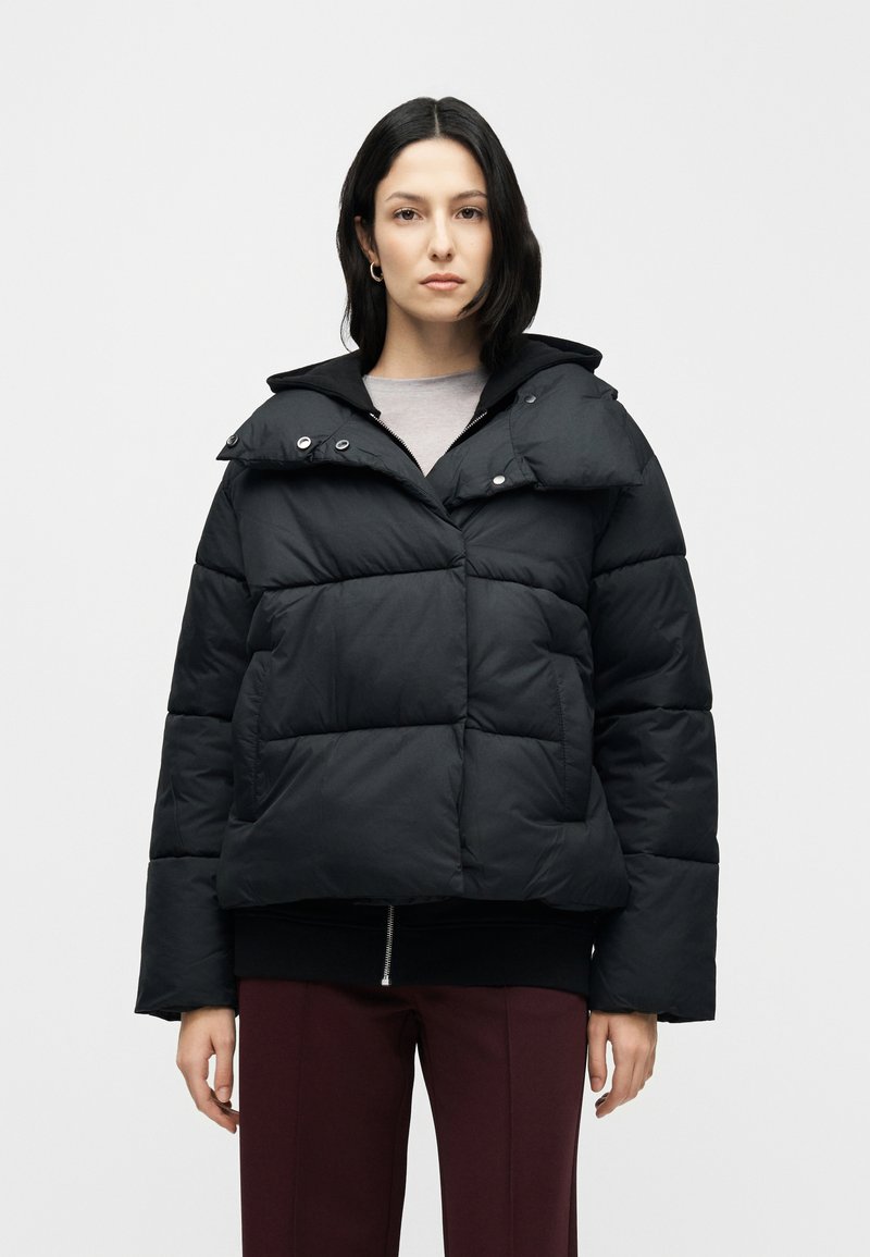 Schwarze Steppjacke mit Kapuze, mit gestepptem Design, Druckknöpfen und kurzem Schnitt, kombiniert mit einem grauen Oberteil und burgunderroten Hosen.