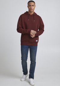 Burgundi värvi kapuutsiga shirt, vaba lõikega, ribidega mansetid, kantud siniste teksade ja valgete tossudega, helehallil taustal.