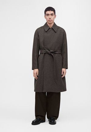 Uomo che indossa un soprabito marrone scuro con cintura, pantaloni scuri a gamba larga e scarpe nere, in piedi davanti a uno sfondo chiaro e uniforme.