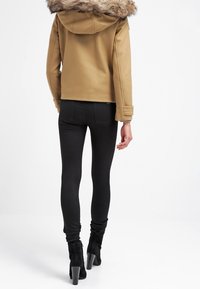 Vrouw in een bruine jas met bontkap, zwart skinny jeans en zwarte enkellaarsjes met hak, staand met haar gezicht naar voren tegen een witte achtergrond.