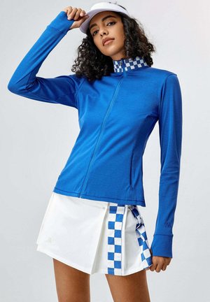 Maglietta atletica blu a maniche lunghe con zip frontale, caratterizzata da un accento a scacchi sul colletto. Abbinata a una gonna bianca con scacchi blu.