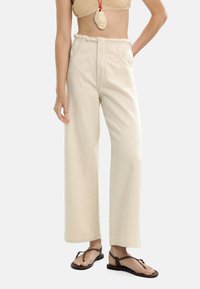 Szerokie, kremowe spodnie z frędzlami w pasie, wykonane z denimu. Posiadają dwie przednie kieszenie oraz luźny krój.