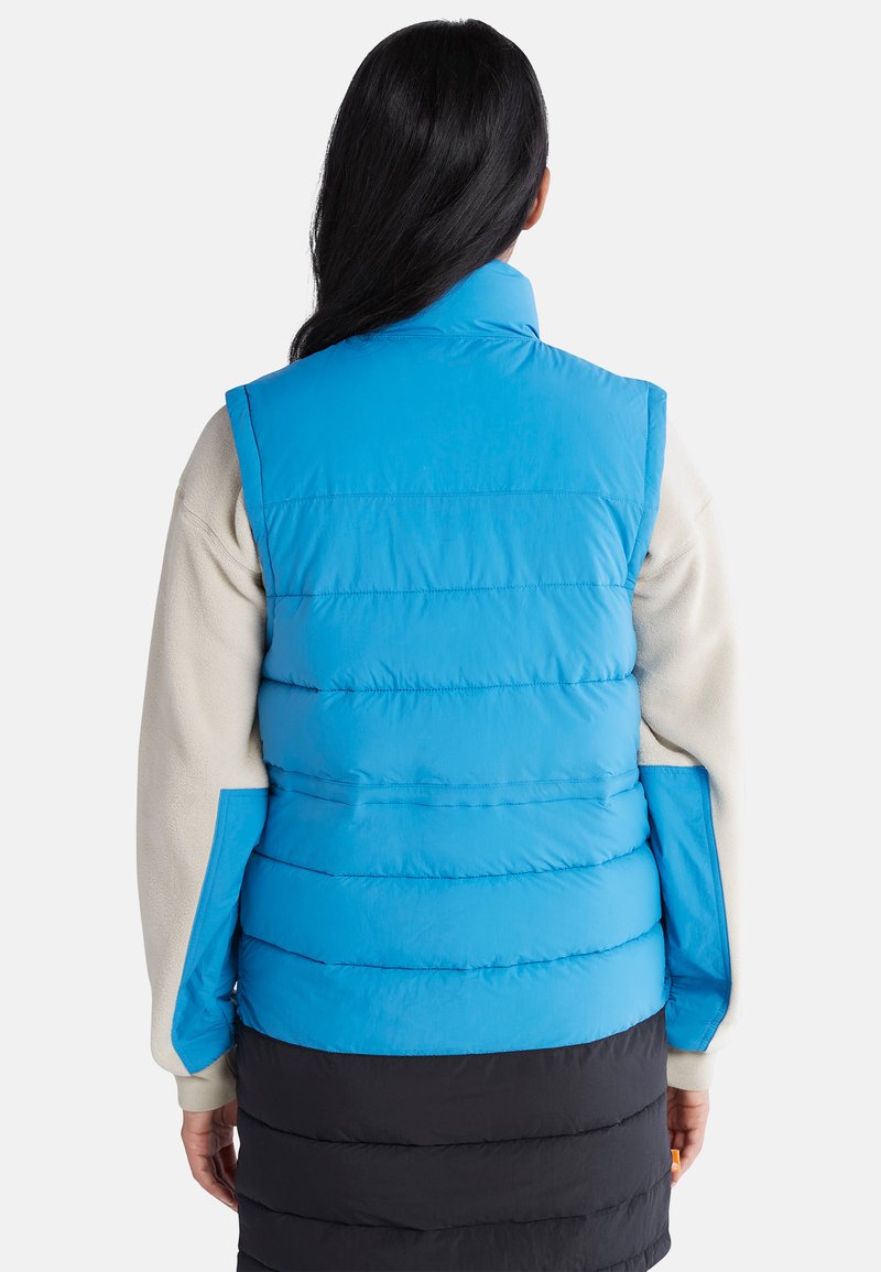 Blaues, puffy Weste mit hohem Kragen, gestepptem Design und glatter Textur. Sie wird über einem beigen Fleece-Oberteil und einem schwarzen Rock getragen.