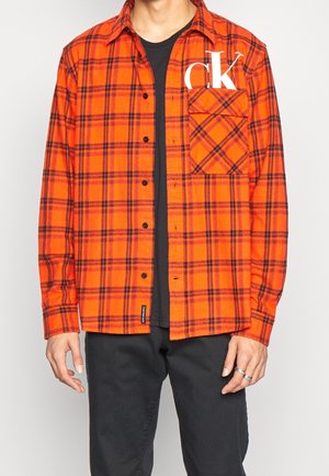 Homme portant une chemise à carreaux orange et noire déboutonnée avec un logo blanc "ck" sur la poche poitrine, par-dessus un t-shirt noir et un pantalon noir.