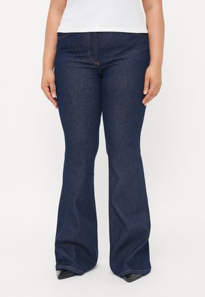 Jean en denim bleu foncé évasé avec une texture lisse, présentant des coutures contrastées et une taille haute, associés à des chaussures noires pointues.