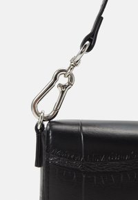 Zbliżenie na srebrny karabinek, który łączy czarny skórzany pasek torby z metalowym pierścieniem torby.