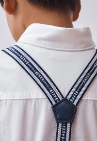Navy and white suspenders feature woven straps with repeating "BAKER" text, a hexagonal leather connector, and are worn over a white shirt.