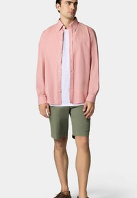 Chemise boutonnée rose clair à manches longues, portée par-dessus un t-shirt blanc, associée à un short vert olive et des chaussures marron.