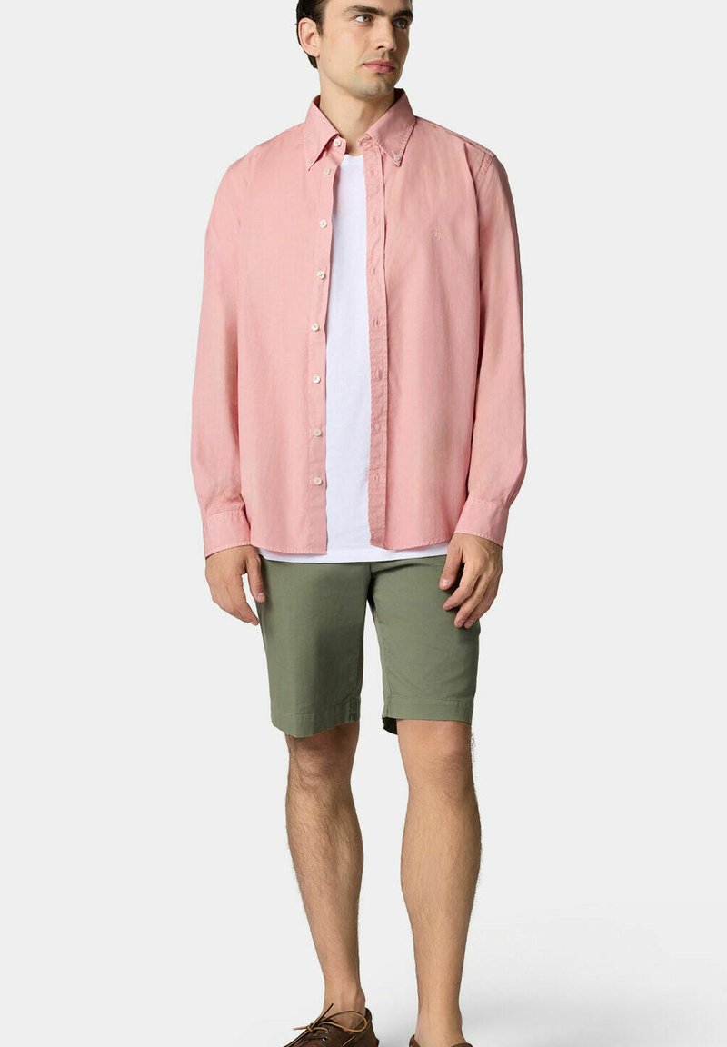 Chemise boutonnée rose clair à manches longues, portée par-dessus un t-shirt blanc, associée à un short vert olive et des chaussures marron.