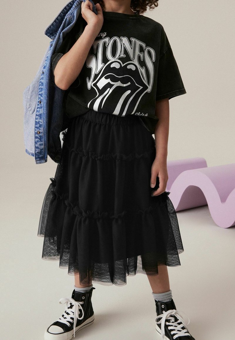Enfant portant un t-shirt noir des Rolling Stones, une jupe en tulle noire à plusieurs couches, des baskets montantes noires, et tenant une veste en jean bleue sur une épaule.