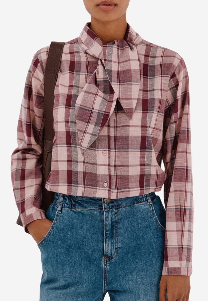 Kostkovaná bavlněná blůza v odstínech růžové a burgundské, s límcem na mašli, zapínáním na knoflíky a dlouhými rukávy. Doplňena modrými džíny.