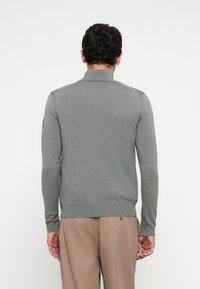 Man wearing a gray long-sleeve sweater and beige pants, standing facing away against a plain white background.