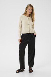 Cardigan en tricot de couleur crème avec un col rond et une fermeture à boutons, associé à un pantalon noir, ample, avec cordon de serrage. Sandales plates.