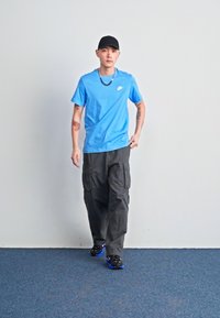 Modra bombažna majica s logojem, črne cargo hlače in črne teniske z modrimi detajli. Figurica stoji na modri preprogi ob svetli steni.