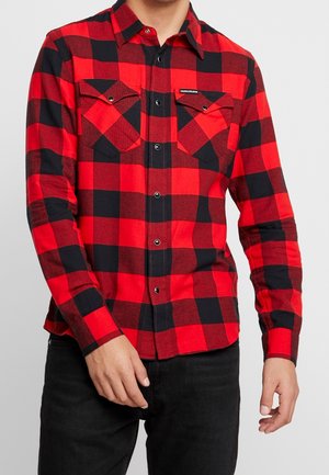 Homme portant une chemise à manches longues à carreaux rouge et noir avec deux poches poitrine, associée à un pantalon noir.