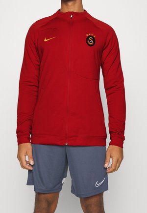 Personne portant une veste rouge zippée avec un logo sportif et un short gris Nike sur un fond blanc uni.