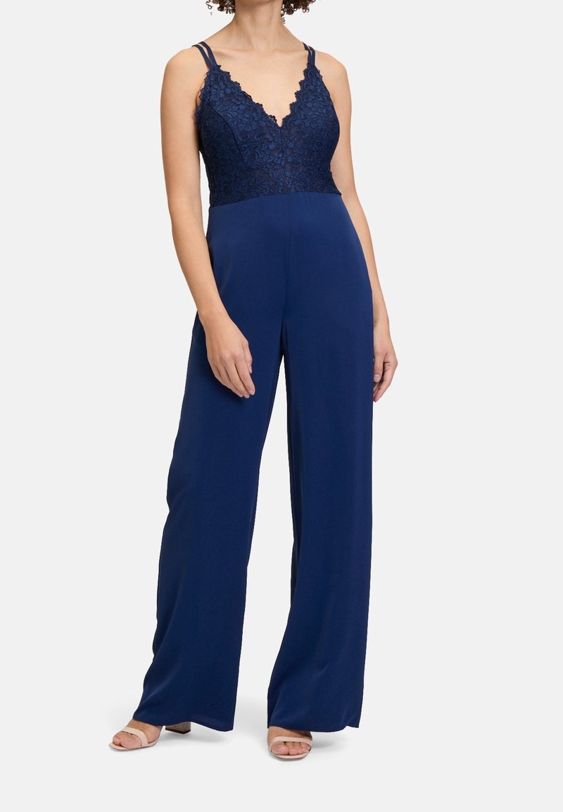 Femme portant une combinaison sans manches bleu marine avec corsage en dentelle à encolure en V et pantalon à jambes larges, associée à des sandales ouvertes de couleur claire.
