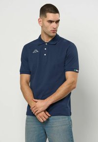 Rövid hajú férfi, sötétkék háromgombos galléros pólót és farmert visel, oldalra néz, kezeit összekulcsolja maga előtt.