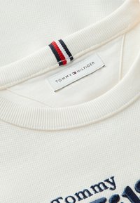 Gros plan d'un sweat-shirt blanc texturé à col rond avec étiquette Tommy Hilfiger et boucle en tissu rouge, blanc et bleu à l'intérieur du col.