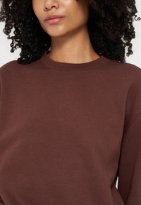 Femme aux cheveux bouclés et foncés portant un sweat-shirt marron uni à col rond, montrée des épaules aux lèvres sur un fond clair.