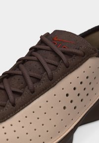 Καφέ και μπεζ αθλητικό παπούτσι Nike με περατό πλαϊνό πάνελ, καφέ κορδόνια και κεντημένο πορτοκαλί λογότυπο Nike στη γλώσσα.