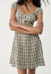 Brązowa i beżowa sukienka w kratę z bufiastymi rękawami, kwadratowym dekoltem i zapięciem na guziki. Posiada warstwową spódnicę z falbaną na dole.