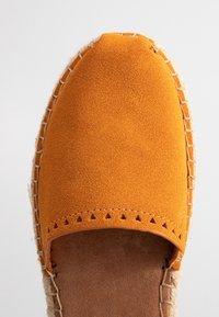 Chaussure espadrille en daim orange avec semelle en jute tressée et détails de découpes triangulaires près de l’ouverture, vue de dessus sur fond blanc.