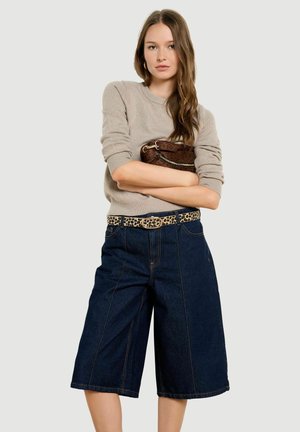 Vrouw die een beige trui draagt, donkerdere wijde denimshorts, een leopardprint riem, en een kleine bruine tas over haar borst houdt.
