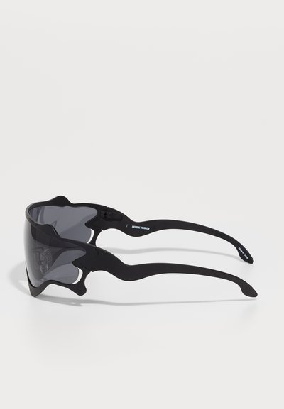 Des lunettes de soleil noires au design élégant et englobant, avec des verres foncés et des montures de forme unique pour un look moderne.