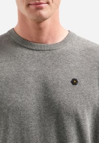 Man in een grijze gebreide trui met een klein zwart hexagonaal logo met gele strepen op de linkerborst.