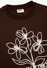 Brauner Sweatshirt mit einem weißen floralen Grafikdesign. Hergestellt aus weichem Material mit einem gerippten Rundhalsausschnitt und langen Ärmeln.